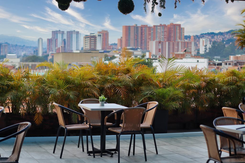 Terraza del hotel con mesas y sillas entre palmeras y vista a los edificios de Bello.